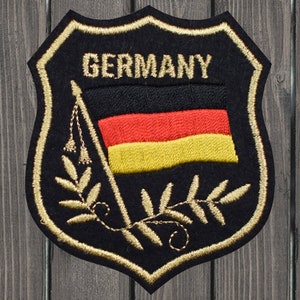 Peut inclure: Un écusson en feutre noir avec une bordure brodée dorée en forme de bouclier. L'écusson présente un drapeau allemand brodé en or avec le mot "GERMANY" au-dessus. Le drapeau est entouré d'une couronne brodée en or.