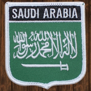 Pode incluir: Um remendo bordado verde com costuras brancas. O remendo apresenta a bandeira da Arábia Saudita, com a escrita árabe e uma espada. O texto "SAUDI ARABIA" está impresso em branco sobre um fundo preto na parte superior do remendo.