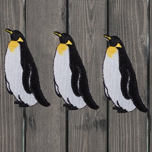 Può includere: Tre toppe ricamate di pinguini in bianco e nero con becchi e piedi gialli. Le toppe sono su uno sfondo di legno.