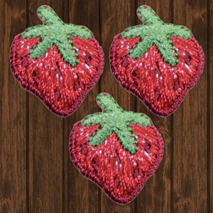 May include: Three red embroidered strawberry patches with green leaves. The patches are on a brown wooden background.