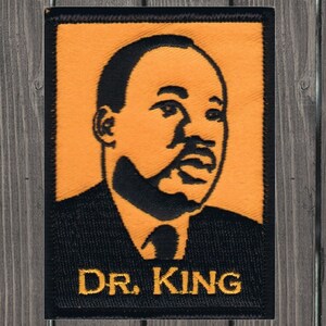 Puede incluir: Parche bordado en negro y naranja con un retrato del Dr. Martin Luther King Jr. con el texto "DR. KING" debajo.