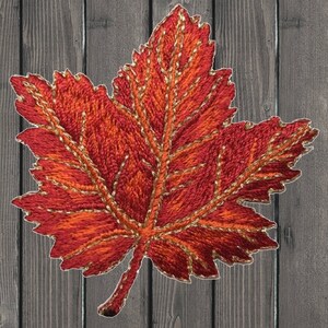 May include: A red and orange embroidered maple leaf with gold thread outlining the veins and edges. The leaf is on a wood background.
