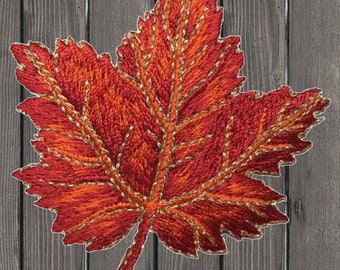 Può includere: Una foglia d'acero ricamata in rosso e arancione, con filo dorato che delinea le venature e i bordi. La foglia è su uno sfondo di legno.