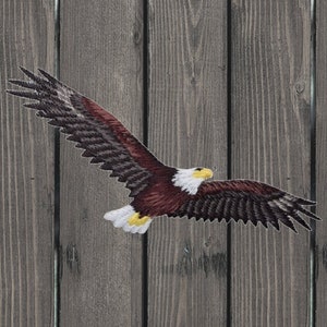 Könnte beinhalten: Gestickter Aufnäher eines fliegenden Weißkopfseeadlers. Der Adler hat einen weißen Kopf, einen gelben Schnabel und braune und weiße Flügel. Der Aufnäher befindet sich vor einem verwitterten Holzgrund.