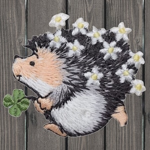 May include: Embroidered patch of a cute hedgehog with a white and black body, holding a green four-leaf clover. The hedgehog has a white face and is wearing a crown of white flowers.