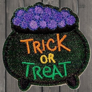 Puede incluir: Un parche temático de Halloween con un caldero verde con las palabras "TRICK OR TREAT" en naranja y verde. El caldero tiene una sustancia morada y burbujeante en su interior. El parche tiene una textura brillante.