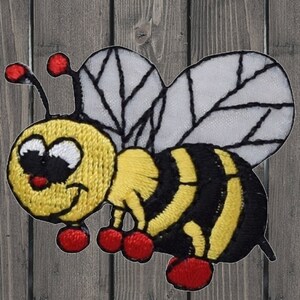 May include: A yellow and black embroidered bee with red accents and white wings. The bee has a cartoon-like face with big eyes and a smile.