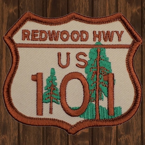 Può includere: Una toppa ricamata marrone e bianca con il testo "Redwood Hwy US 101" e due alberi verdi.