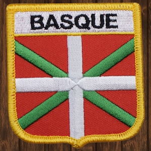 Peut inclure: Un écusson brodé rouge, blanc et vert avec le mot "BASQUE" en lettres noires sur fond blanc. L'écusson a une bordure jaune et un fond pointu.