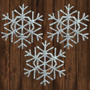 May include: Three silver glitter snowflakes on a brown wood background.