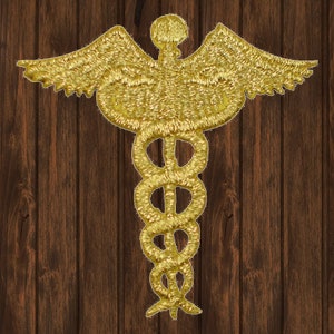 May include: Gold embroidered medical symbol with a snake wrapped around a staff and wings. The symbol is on a brown wooden background.