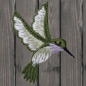 Puede incluir: Parche bordado de colibrí verde y blanco con la garganta morada. El parche es perfecto para agregar un toque de naturaleza a tus proyectos.
