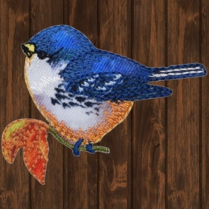 May include: Embroidered bluebird with white and orange accents perched on a branch with a red leaf. The bird is facing right and has a yellow beak.