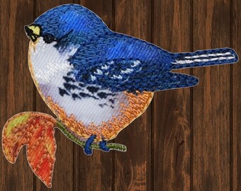Può includere: Un passero blu ricamato con accenti bianchi e arancioni appollaiato su un ramo con una foglia rossa. L'uccello è rivolto a destra e ha un becco giallo.