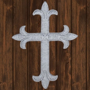May include: A silver-coloured, ornate cross with a fleur-de-lis design on a brown wooden background.