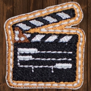 Può includere: Una clapperboard ricamata in bianco e nero con un bordo dorato. La clapperboard ha tre linee bianche e una sezione a strisce bianche e nere nella parte superiore.