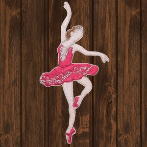 Può includere: Toppa ricamata di ballerina rosa con un tutù bianco e dettagli argentati. La ballerina è in una posa di danza con una gamba sollevata.