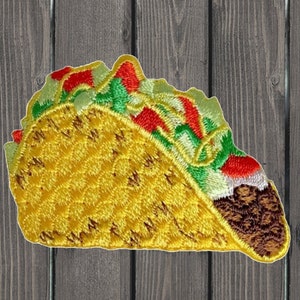 May include: A yellow embroidered taco with red, green, and white fillings. The taco is on a wooden background.