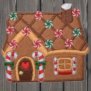 Op de afbeelding: Een bruin peperkoekhuis-patch met rode, witte en groene snoepaccenten. Het huis heeft een hartvormige deur, zuurstokaccenten en een schoorsteen. Het dak is versierd met rode en groene snoepjes.