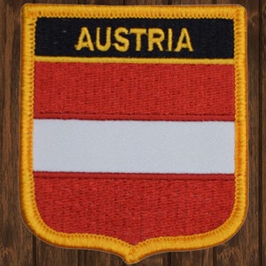 Könnte beinhalten: Ein roter, weißer und schwarzer gestickter Aufnäher mit dem Wort "AUSTRIA" in gelben Buchstaben. Der Aufnäher hat die Form eines Schildes mit einem goldenen Rand.