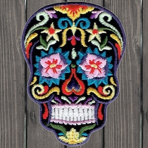 May include: A colorful embroidered sugar skull patch with intricate floral designs in shades of pink, blue, yellow, and red. The skull is black with white teeth and a purple outline.