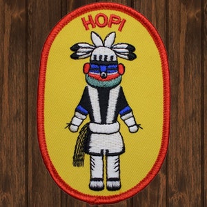 May include: A yellow oval embroidered patch with a red border. The patch features a stylized figure with a black and white striped body, a blue and red face, and a green headdress. The word "HOPI" is embroidered in red letters at the top of the patch.