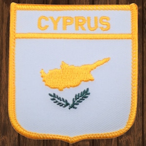 May include: Embroidered patch featuring the word "CYPRUS" in gold above a gold outline of the island of Cyprus. Below the island is a green olive branch design. The patch has a white background with a gold border.