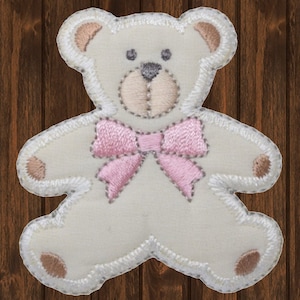 Könnte beinhalten: Ein gestickter Aufnäher in Form eines Teddybären. Der Bär ist cremefarben mit hellbraunen Akzenten an Ohren, Pfoten und Schnauze. Eine rosa Schleife ist auf die Brust genäht. Der Aufnäher hat einen weißen, gestickten Rand.
