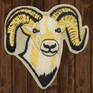May include: Embroidered patch of a white and yellow bighorn sheep with black horns. The sheep is facing forward with its head tilted slightly down. The background is a light beige.