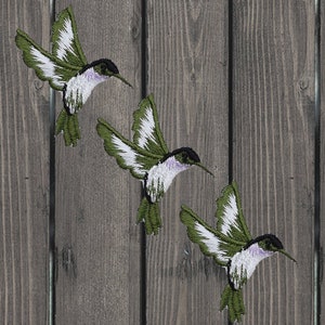 May include: Three green and white embroidered hummingbird patches on a wooden background.