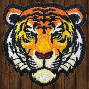 May include: Close-up of an embroidered tiger face patch. The tiger has orange and black stripes, a white muzzle, and a pink nose.