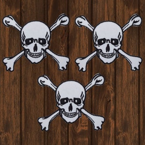 May include: Three white skull and crossbones patches on a brown wooden background. The patches are black and white and have a simple design.