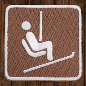 May include: A brown fabric patch with a white outline. The patch features a white silhouette of a person sitting in a ski lift chair with a ski slope underneath.