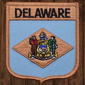 以下が含まれることがあります： 茶色と青色の刺繍パッチで、上部に黒い文字で「DELAWARE」と書いてあり、中央にはデラウェア州の州章があります。州章は菱形で、茶色の背景に、黄色と青色の盾があり、馬と農夫が描かれています。