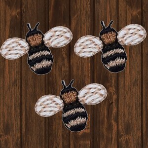 Puede incluir: Tres parches bordados de abejas con rayas blancas y negras, cuerpo marrón y alas blancas. Las abejas están sobre un fondo de madera marrón.