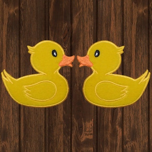 May include: Two yellow duck patches with orange beaks and black eyes are displayed against a brown wooden background. The ducks face each other, suggesting a playful or decorative theme. The patches are likely for clothing or craft projects.