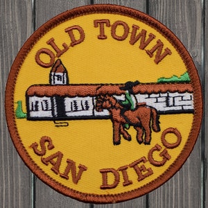May include: A yellow embroidered patch with brown stitching. The patch features a white building with a brown roof, a brown horse with a rider, and the text "Old Town San Diego".