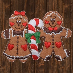 Może przedstawiać: Para gingerbreadów trzymających laskę cukrową z zieloną kokardką. Ludziki z piernika są brązowe z białą polewą i czerwonymi sercami.