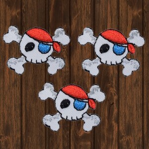 May include: Three white and gray embroidered pirate skull patches with red and blue bandanas. The patches are on a brown wooden background.