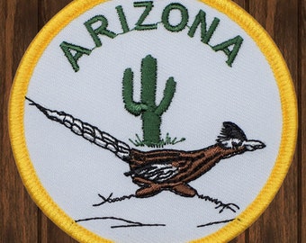 Puede incluir: Parche bordado con la palabra "ARIZONA" en verde sobre un cactus verde y un correcaminos marrón y blanco. El parche tiene un fondo blanco con un borde amarillo.