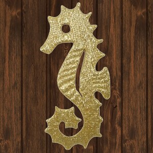 May include: A golden seahorse silhouette with a textured surface. The seahorse is set against a dark brown, vertical wood grain background. The seahorse has a circular eye and spiky protrusions along its back and tail.