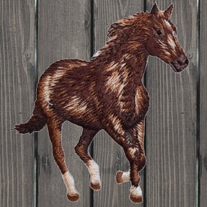 May include: Brown and white embroidered horse patch. The horse is running with its head turned to the side. The patch is on a wooden background.