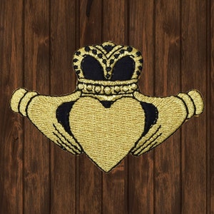 May include: Embroidered Claddagh patch featuring a gold heart held by two gold hands, topped with a gold and black crown. The patch is on a brown wooden background. The Claddagh symbol represents love, loyalty, and friendship.
