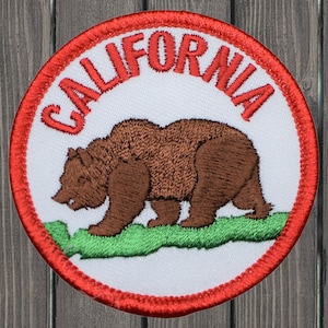 Peut inclure: Un écusson brodé rouge, blanc et vert avec le mot "CALIFORNIA" en lettres rouges autour du bord. Un ours brun marche sur une bande de gazon verte.