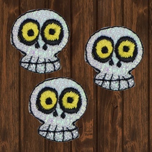 May include: Three white sparkly skull patches with black outlines and yellow eyes. The skulls have a toothy grin.