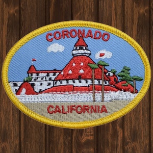 May include: A yellow oval embroidered patch with a red building with a white roof and a white tower. The building is surrounded by palm trees and clouds. The text "CORONADO CALIFORNIA" is embroidered in red on the patch.