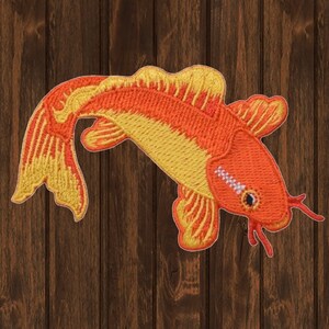 May include: An orange and yellow embroidered iron-on patch of a koi fish. The fish is swimming to the right and has a white stripe on its side.