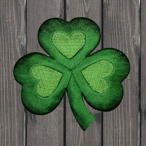 May include: Embroidered shamrock patch featuring a three-leaf clover design. The shamrock is a vibrant green with heart-shaped leaves and a textured appearance. The background is a wooden surface.
