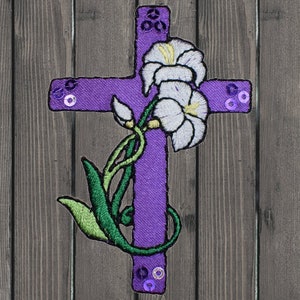 May include: Purple embroidered cross with white flowers and green leaves. The cross is decorated with purple sequins.