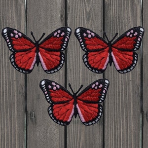 May include: Three red and white embroidered butterfly iron-on patches. The butterflies have black outlines and pink accents.